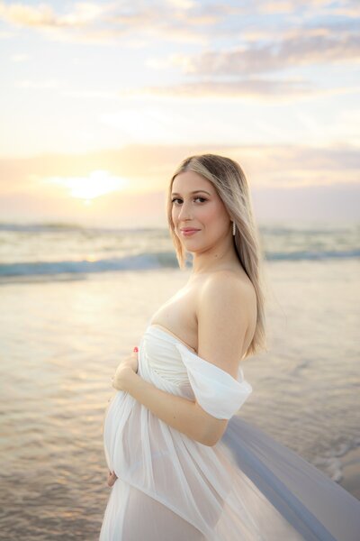 Maternity portrait of mom to be in a white flowing dress during golden hour on Holmes Beach on Anna Maria Island