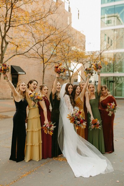 A bride in a white dress and her bridesmaids in assorted earth-tone dresses cheerfully raising their bouquets on a city street during a stress-free Iowa wedding.