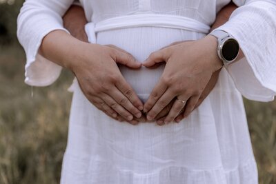 Frau in weissem Kleid haelt Haende Herzfoermig auf ihrem Babybauch