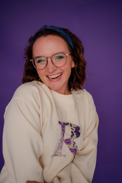 Woman poses for the camera against a purple colored backdrop smiling at the camera.