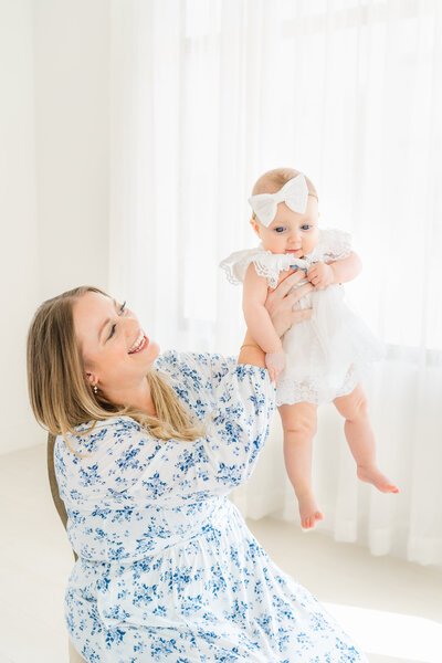 Austin Lifestyle Newborn Photographer | A mother sits on a chair in an all-white studio and holds her baby girl up in the air.