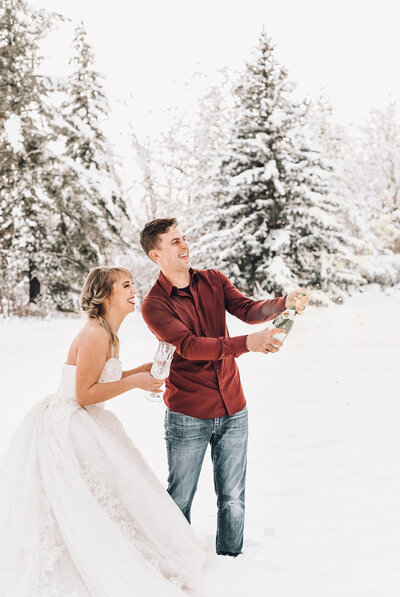 woman in white wedding dress with man in jeans and button down shirt holding champagne glasses outside in the snow