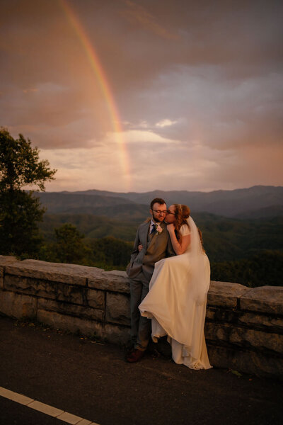 sunset elopement photos with couple sitting on an outlook in the Foothills with a rainbow  behind the Smoky Mountains as the couple embraces and smiles at one another as they elope to Gatlinburg 