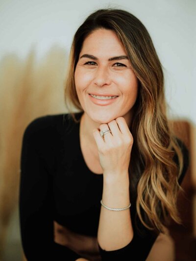 A woman with wavy brown hair wearing a black t-shirt and pants sits on a wooden chair, smiling at the camera. Decorative pampas grass is visible in the background, evoking the warm ambiance of Falmouth Maine Medical Aesthetics.