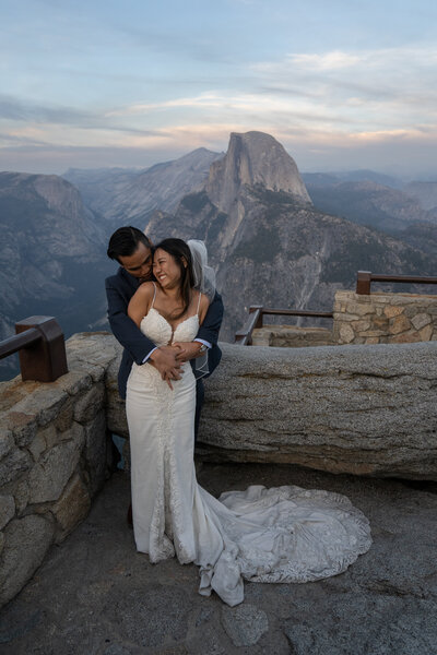 Bride and groom are all smiles while hugging on Glacier Point, with Half Dome and colorful clouds shine in the twilight.