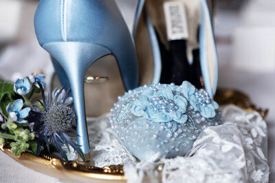 Detail of Badgley Mischka satin blue embelished shoes on a golden tray with adorned with blue petais and a thistle flower.