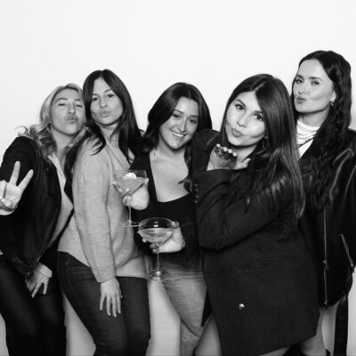 Five women standing close together in front of a white backdrop, holding drinks and making playful poses, including peace signs and blowing kisses toward the camera.