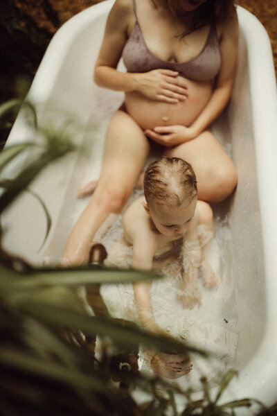 A pregnant mother sitting in a bathtub with her toddler. 