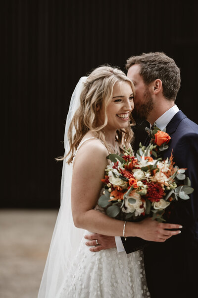 bride and groom holding each other by the waist and holding flowers between them. The bride is smiling while he groom whispers in her ear.