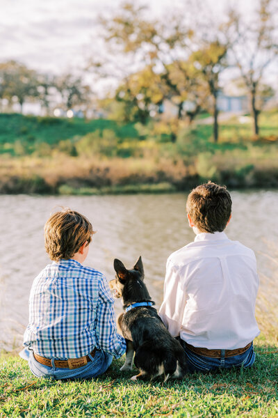 Two boys and their dog sitting on the grass facing the water