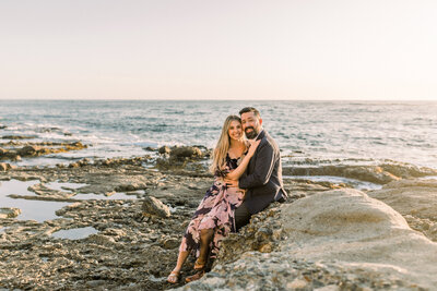 Bride to be sits on her fiance's lap while he sits on a rock at Treasure Island Beach with the ocean behind them