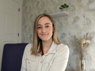 Lexi smiles kindly at the camera, with closed lips. She is wearing a tan, fleece button-down top. Behind her, the wall is covered in a plant-patterned wallpaper, and a small houseplant is visible on a shelf above her head.