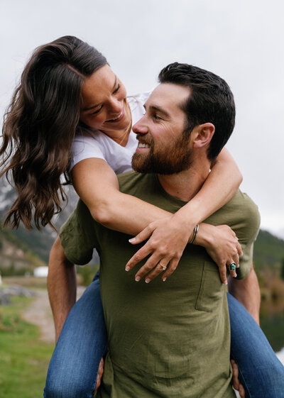 casual engagement session with Trevor giving Jasa a piggyback. Couple is looking at each other and laughing.