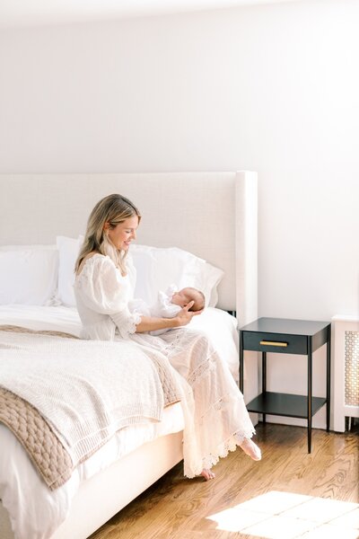 A mother wearing a white dress sits on a bed while holding her newborn in her arms during newborn photos photographed by New Jresey newborn Photographer Kate Voda Photography