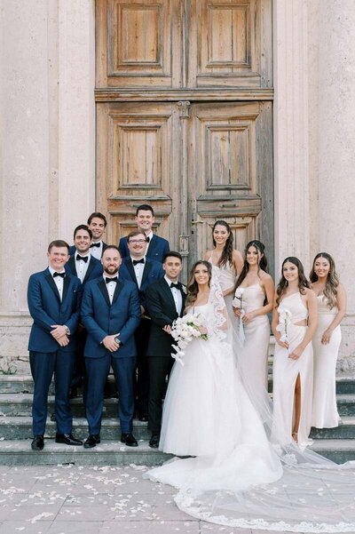 Bride and groom with wedding party in tuxedos and champagne dresses, classic group portrait near historic doors