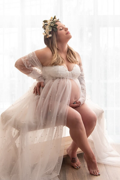 Expecting mother in a white chiffon dress, white curtain background and sitting on a bench