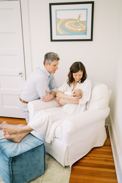 Mom sits in white chair in nursery with newborn baby boy and dad kneeling next to her