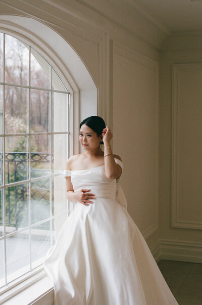 Elegant bride in a flowing gown poses confidently in front of a sunlit window during her wedding portraits