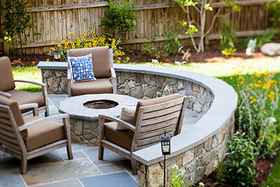 Beautifully completed backyard featuring a curved stone seating area with a built-in fire pit, comfortable patio chairs, and lush landscaping with vibrant flowers.