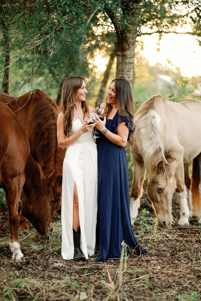two wives sharing a toast of wine in glasses in front of their horses
