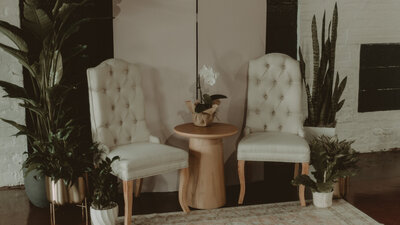 Stunning photoshoot backdrop with 2 vintage, white upholstered accent chairs and a beautiful wooden side table between them, rented from Beautifully Layered Event Rentals in Milwaukee, WI and plants surrounding the setup