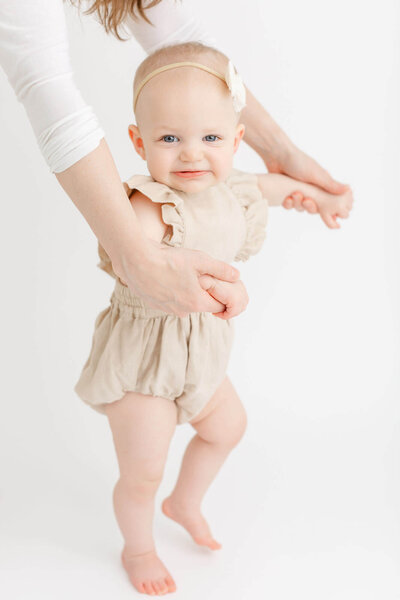 Baby girl in cute neutral colored romper is holding mom's hands while taking a few steps. Baby is smiling towards the camera and only mom's hands are in the image, not her face or body.