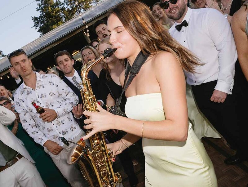 Female saxophone player performing at an outdoor wedding reception surrounded by excited guests.