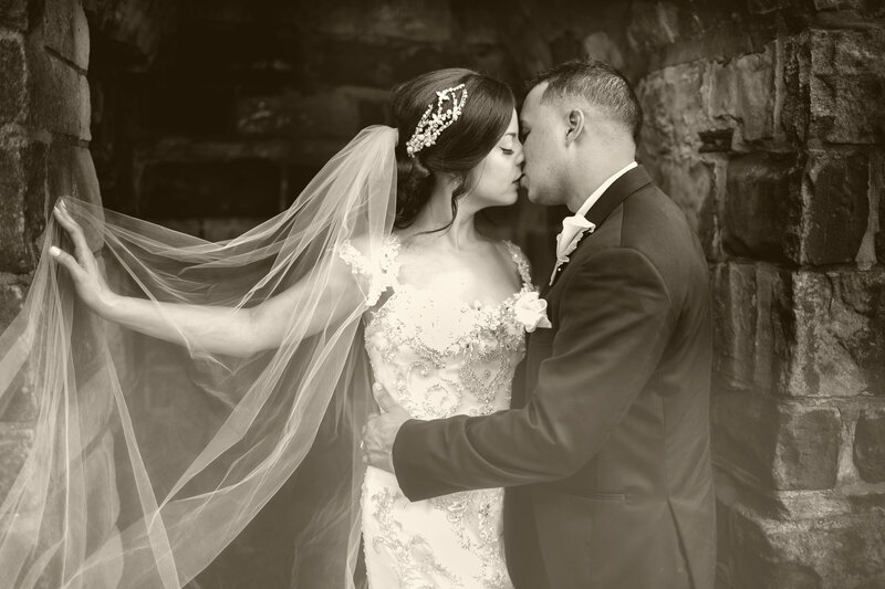 A black-and-white portrait of a bride and groom kissing beside a stone wall, her veil floating behind them