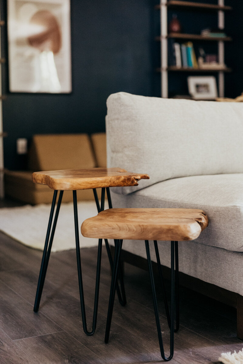 A close up of wooden side tables next to a beige couch with a bookcase out of focus in the background.