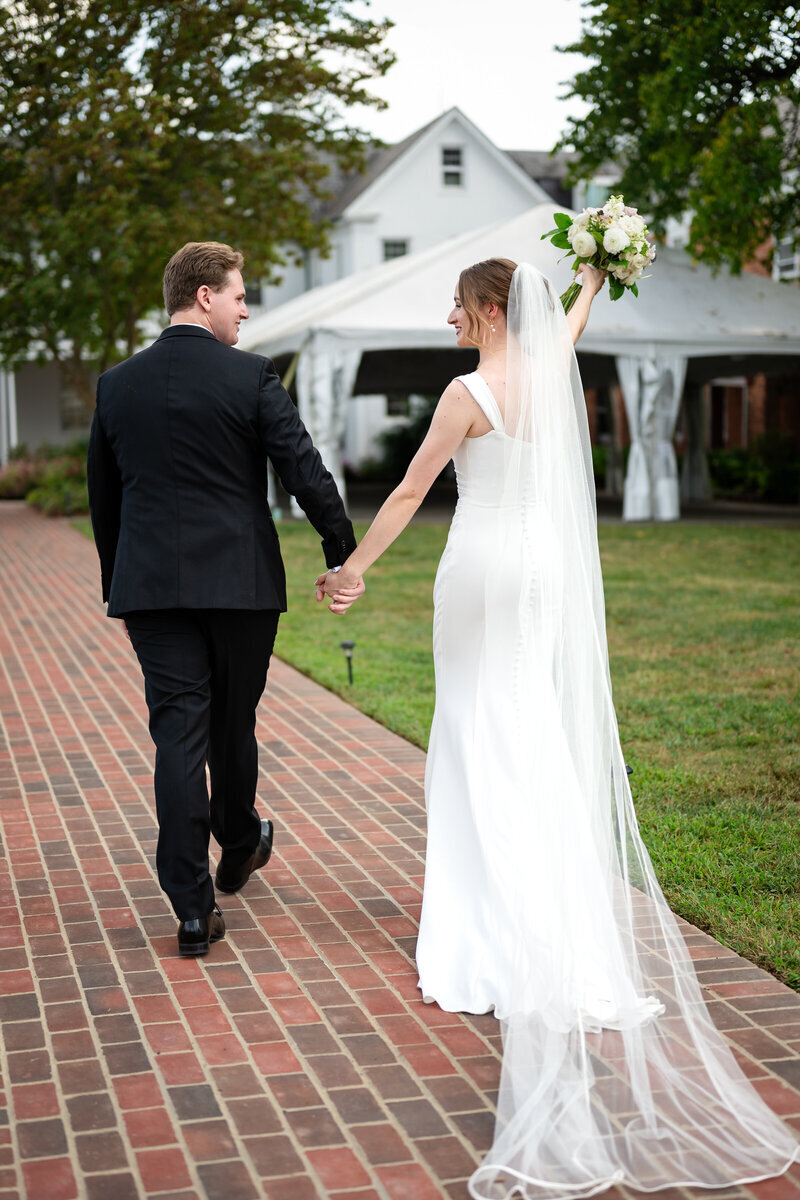 Bride and Groom walk along path while bride holds up bouquet in front of reception area in northern VA