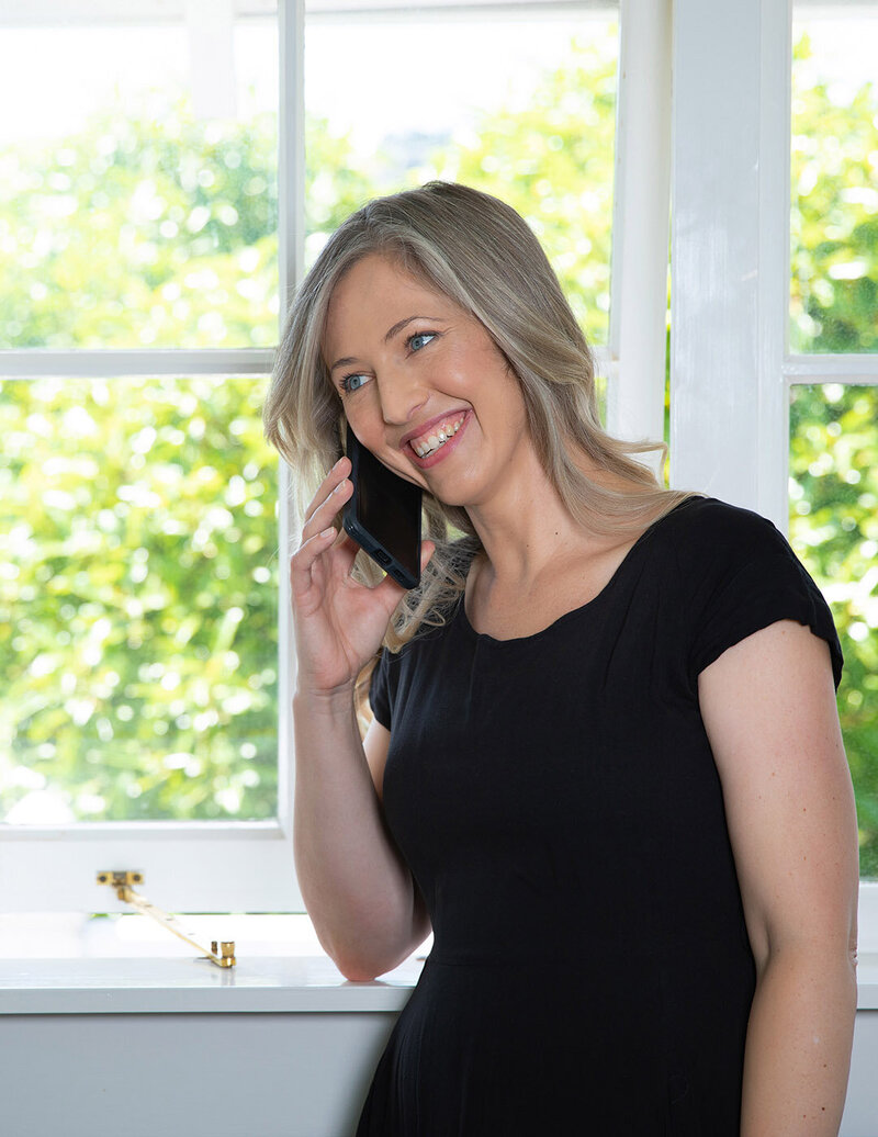 Woman standing by window smiling while talking to client on cellphone