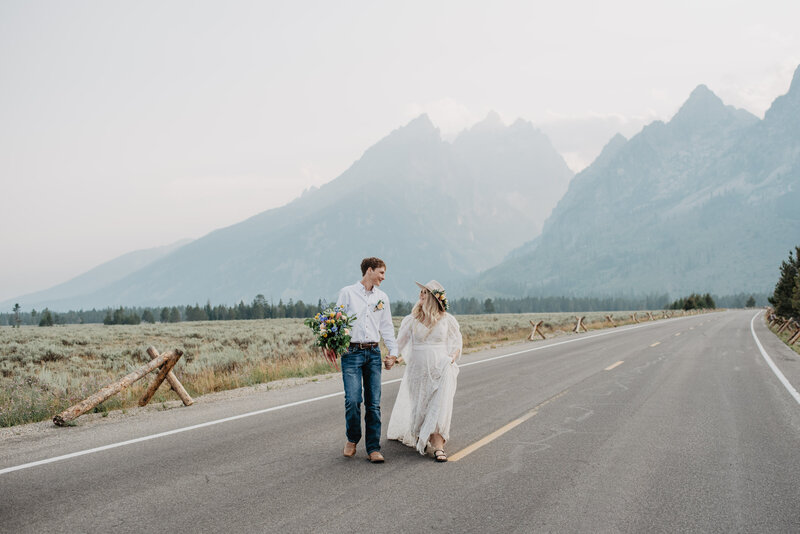 jackson hole wedding with young bride and groom holdin ghands and walking across a back road in the tetons at cathedral turnout for a spring wedding captured by grand teton photographer