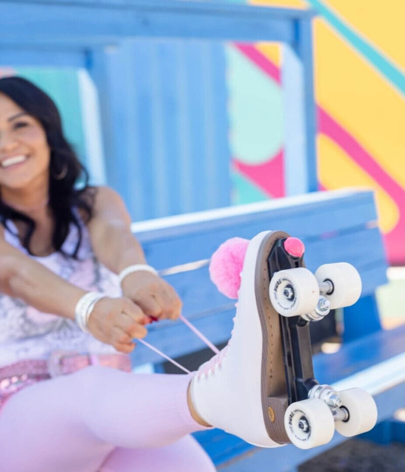 Close-up of a woman adjusting her white roller skate with pink pom-poms, sitting on a blue bench at a colorful roller rink.