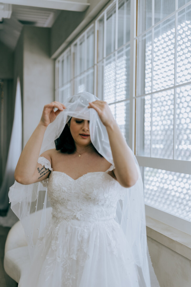 A bride adjusting her veil while standing by a window.