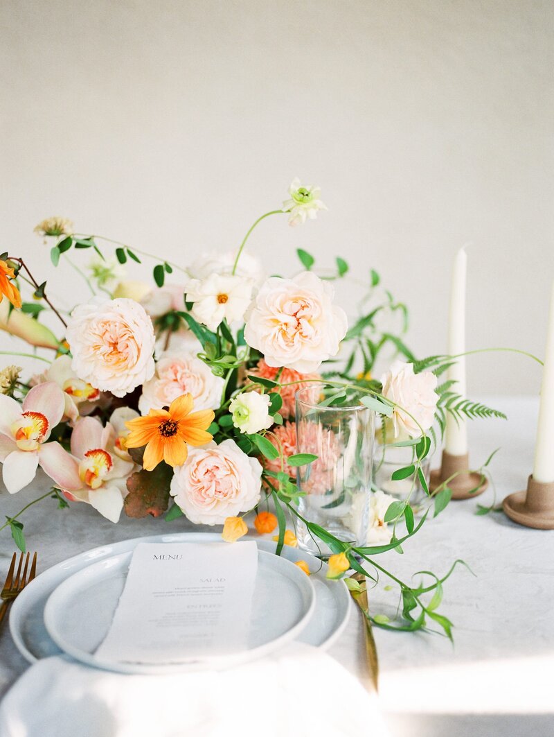 A medium floral arrangement with orange and pink flowers and greenery on a table with a light background