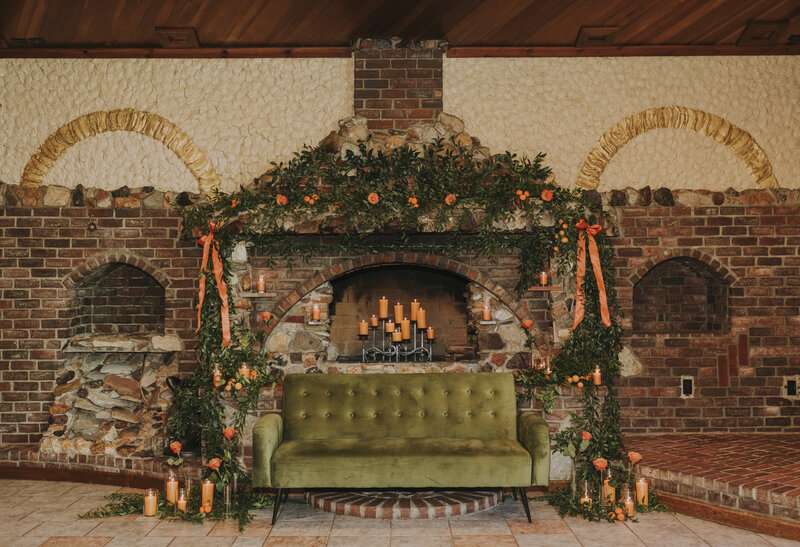 Rustic stone fireplace in the century room at grbic events decorated with greenery, orange flowers, and candles.