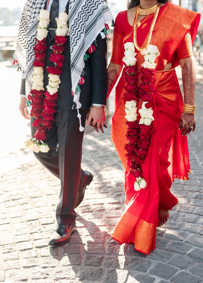 A couple walks hand in hand. The woman wears a red saree with a floral garland, and the man wears a suit with a keffiyeh and garland. Sunlight illuminates the cobblestone path they walk on.