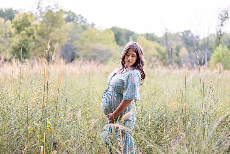 Expecting mother wearing a blue floral dress looking over her shoulder and standing in a field of tall grass. 
