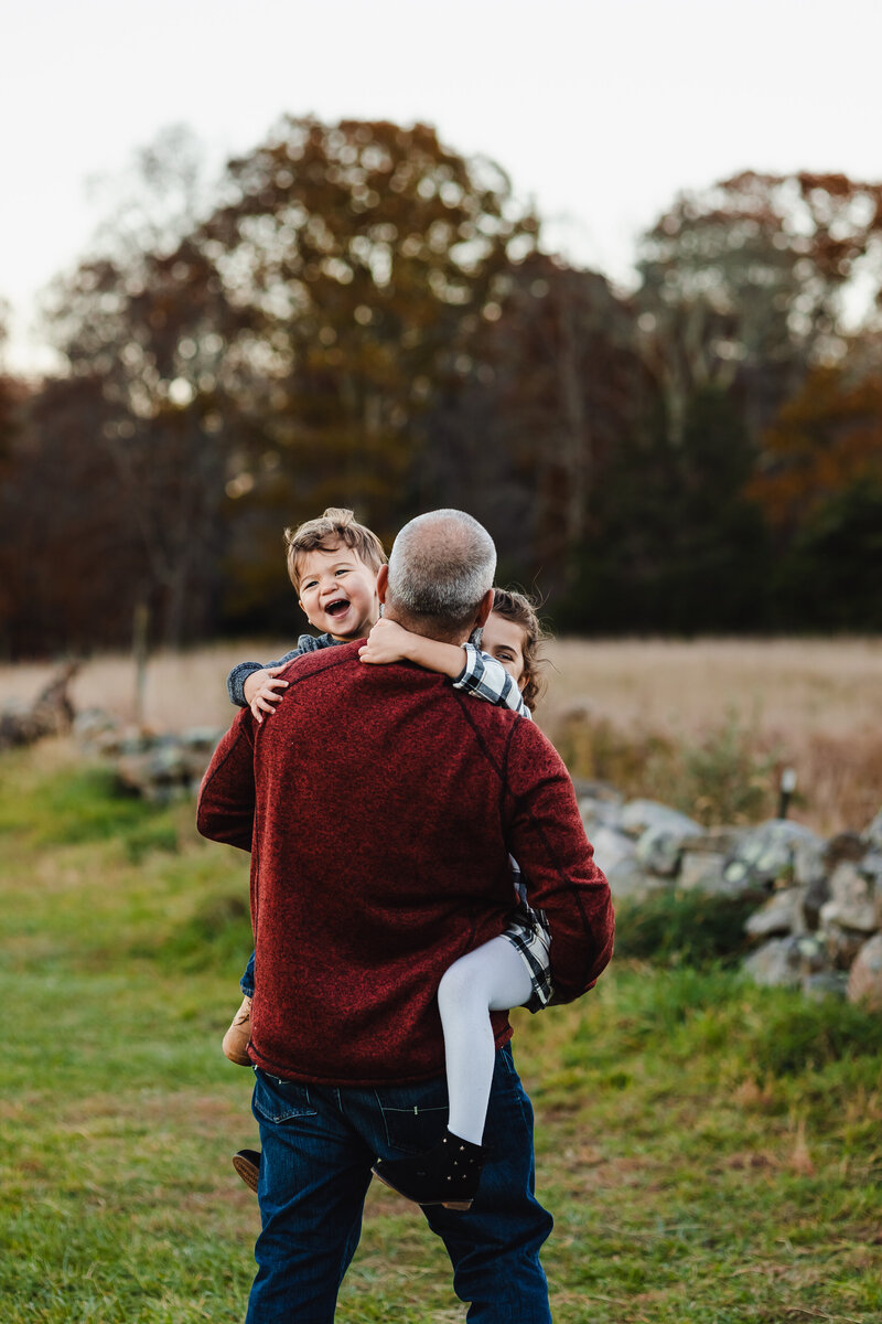 a dad in a red shirt carrying his young smiling son and daughter through a field