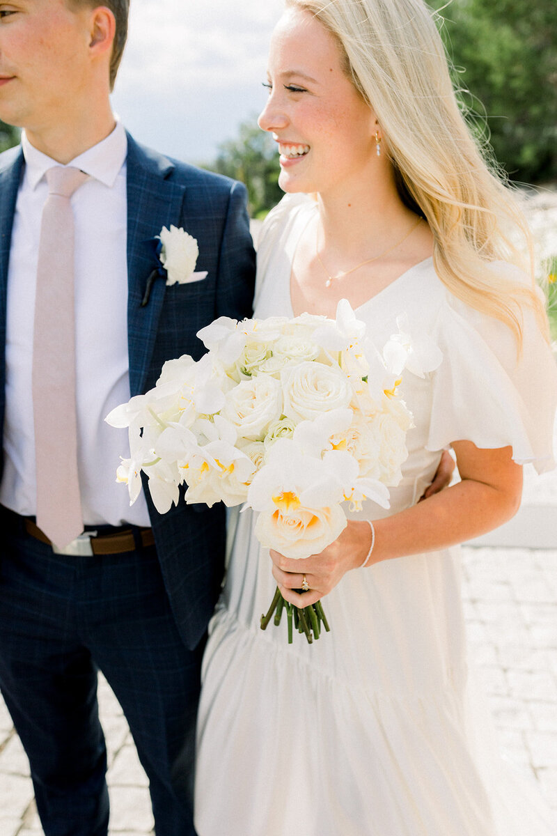 Young, blonde bride holding a bouquet of white flowers next to groom in navy suit.