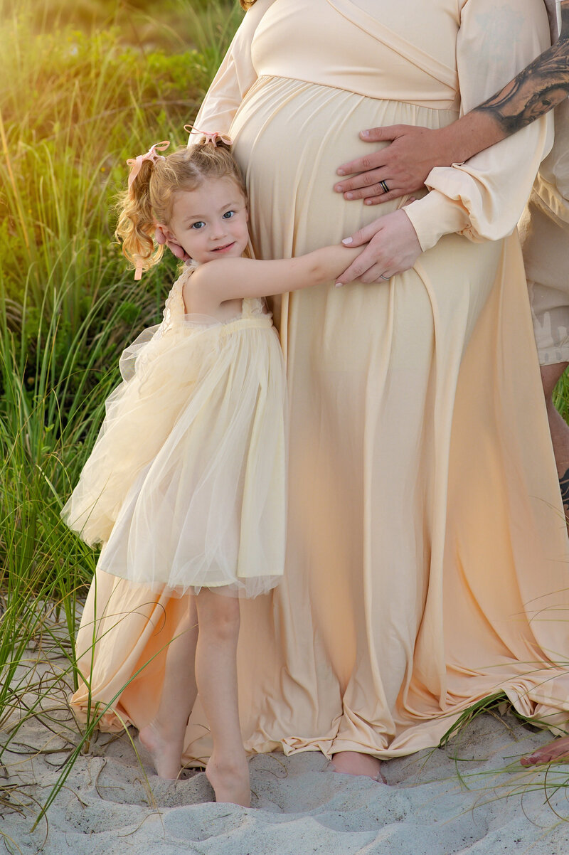 Little girl hugging mom's pregnant belly, both wearing yellow dresses on a beach during a maternity photo session by Orlando photographer Melissa Vinsik
