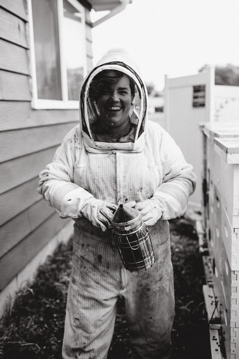A black and white photo of Carrie smiling next to beehives in her bee suit holding a smoker
