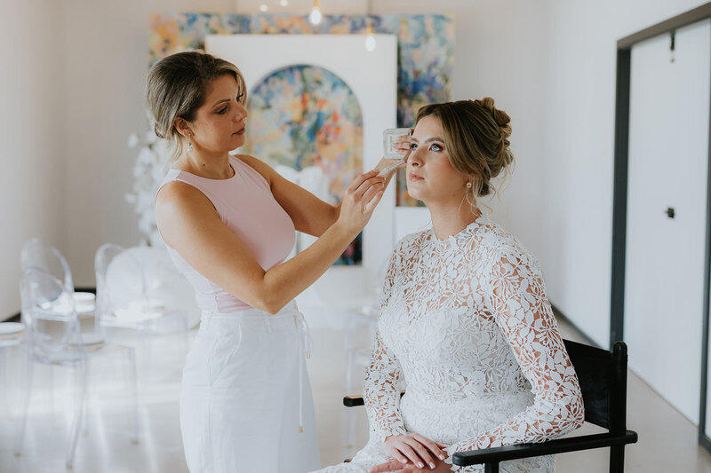 A makeup artist is applying the finishing touches to a bride on her wedding day Elopement in an Art Gallery with a colourful painting behind them, some clear chairs and an arbour. The image is clean and crisp and colourful.