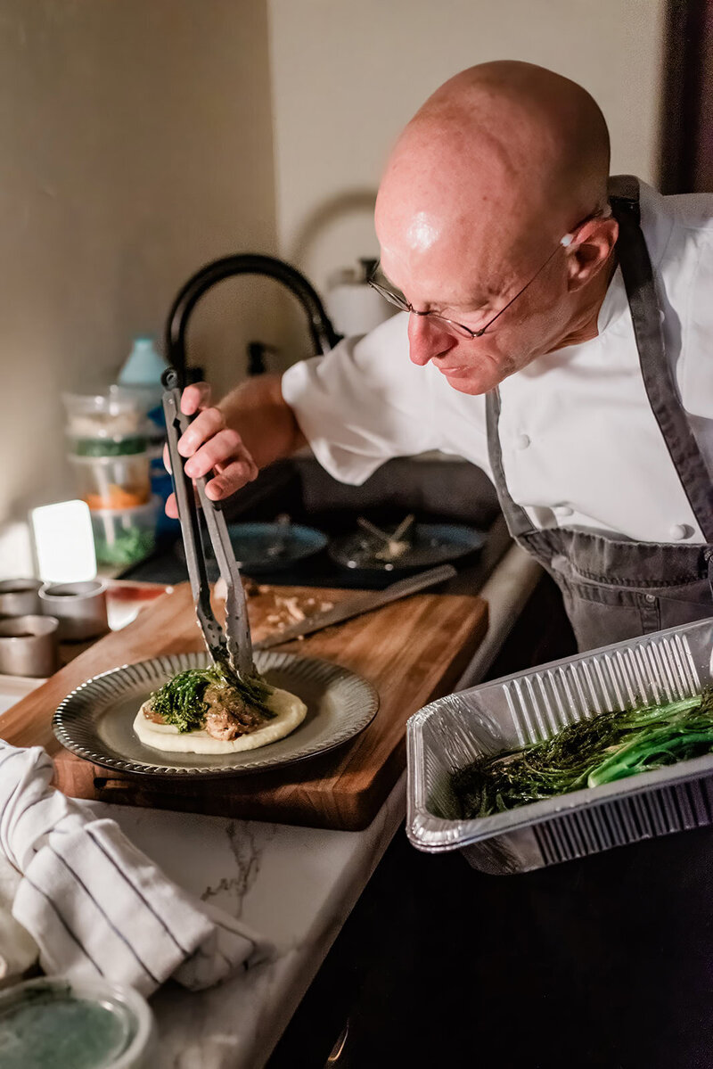 A private chef prepares a bespoke meal at the cabin of the couple the night of their Denali Glacier elopement in Alaska, as part of the adventure luxe elopement package.  He is delicately arranging beautiful vegetables atop the main course on a decorative plate from the sweetheart table.  