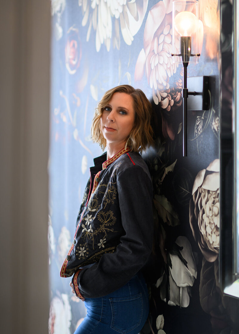 Portrait of Christie Newell standing against a floral mural, representing her bold and artistic photography style.