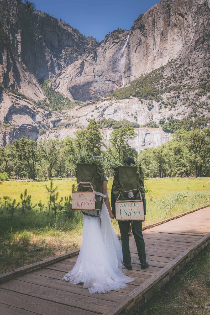 The backs of a bride and groom wearing backpacks with elopement signage during their Yosemite elopement.