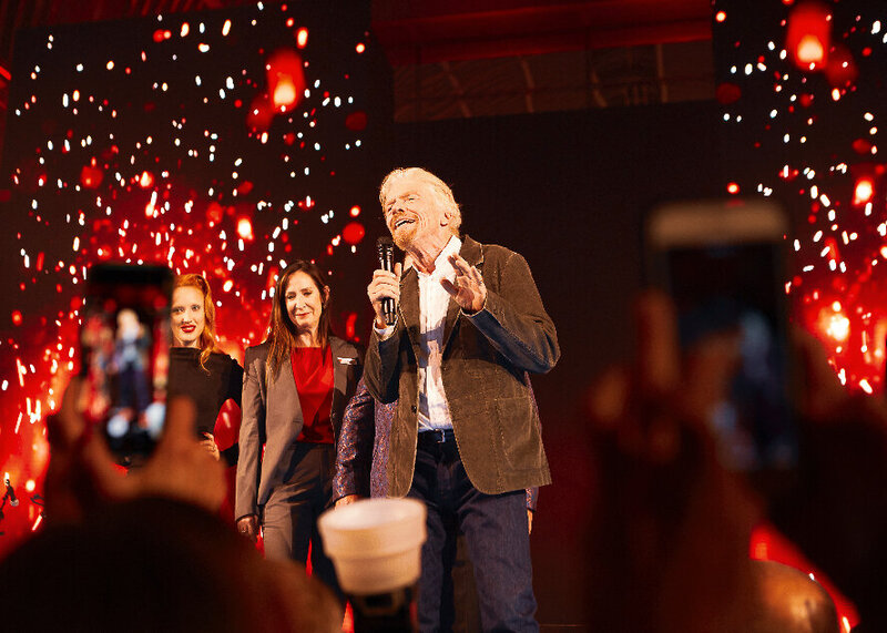 Richard Branson making a speech at a party for Virgin Voyages in London with a red video screen.