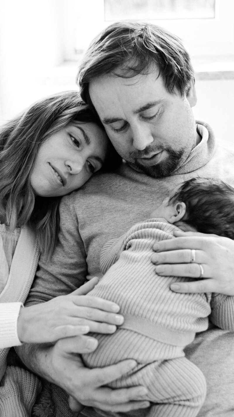 Mother and father holding their newborn closely, heads touching in a quiet, tender moment.