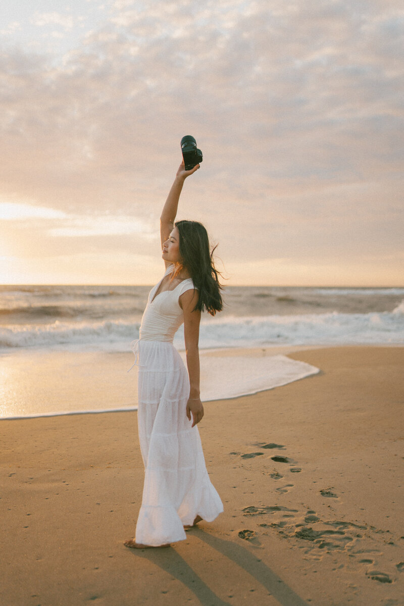 full body photo of Rachel on the beach during sunrise in a white top and maxi skirt raising her arm and holding a camera