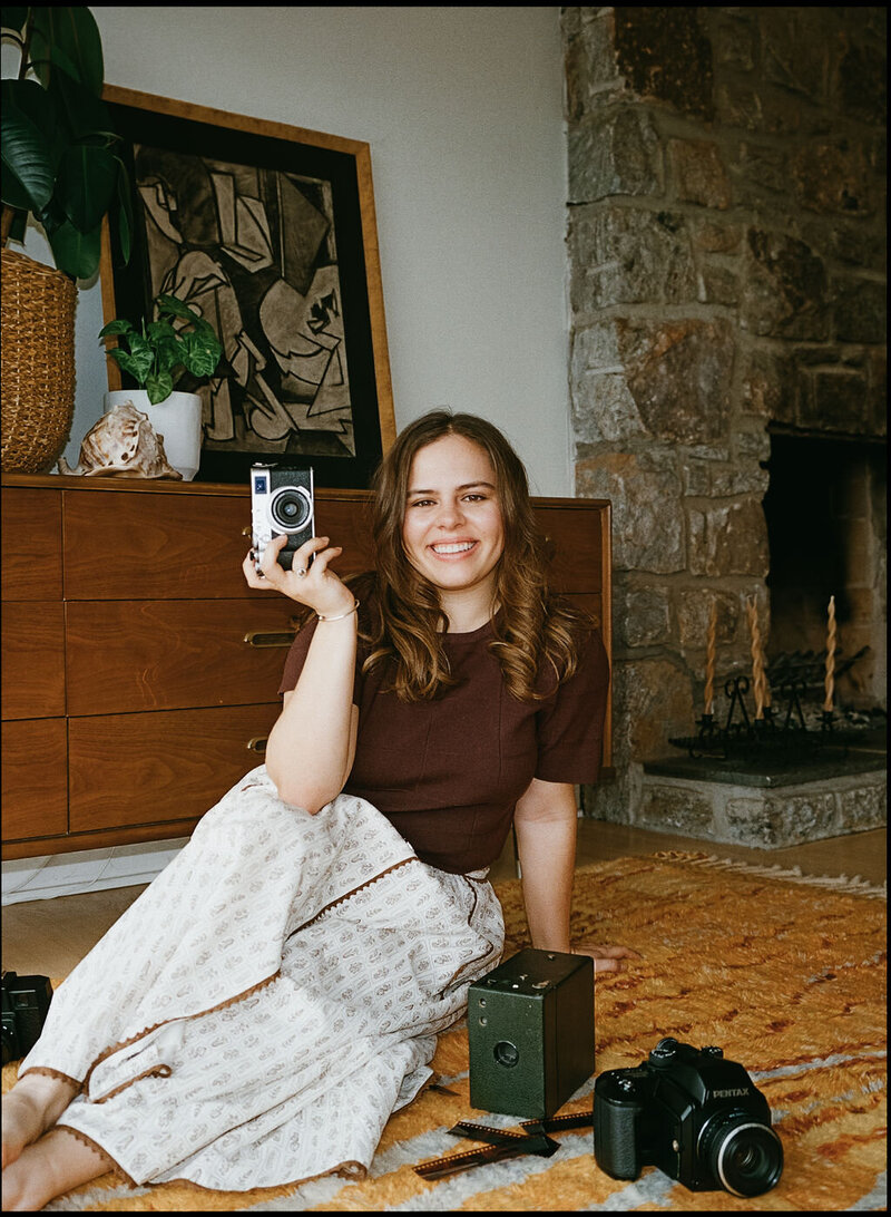 Girl sits smiling with camera on the floor of a house and other film camera are on the ground around her